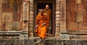 Buddhist monks in orange robes step out of an ancient stone temple in Thailand, showcasing historical architecture.